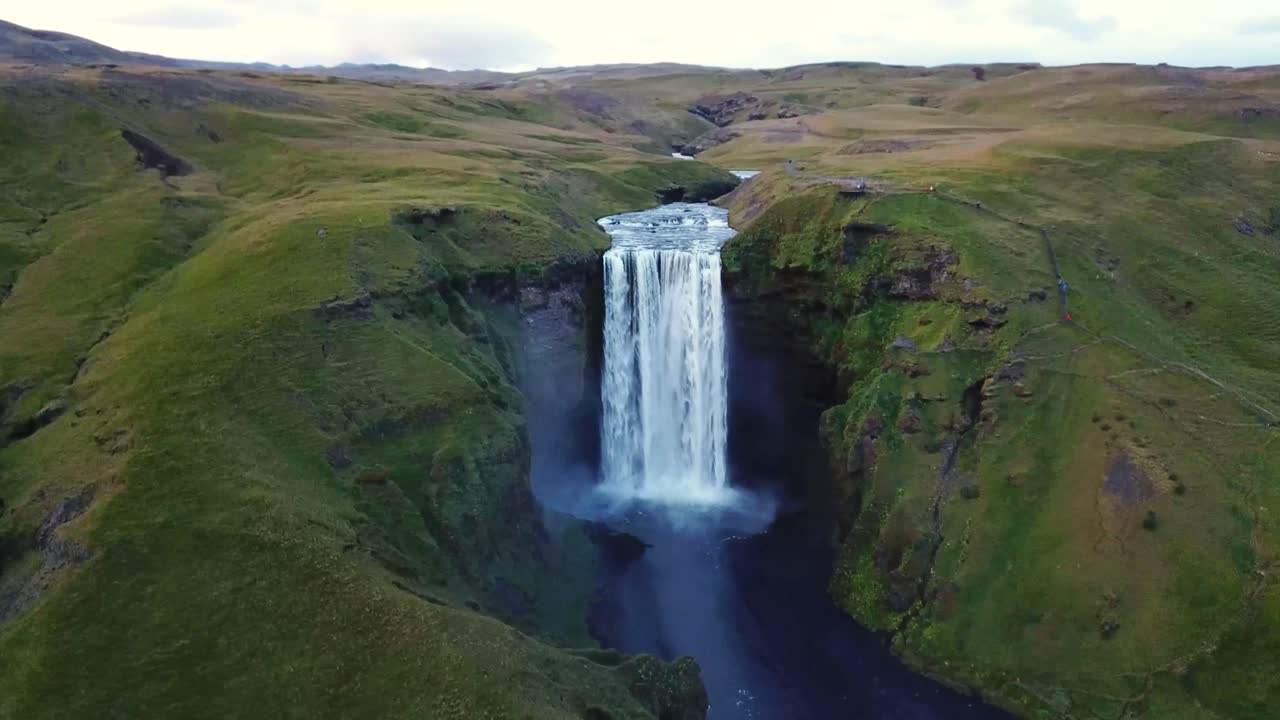 An enchanting aerial view of Skógafoss in Iceland, where a powerful waterfall plunges into a misty basin surrounded by lush green cliffs, winding rivers, and the untouched beauty of Nordic landscapes