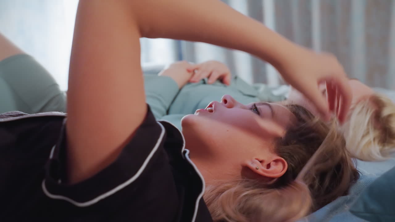 Close up of two girls lying on bed in relaxed indoor atmosphere, one with arm raised holding object possibly phone, other lady resting, with natural light coming through curtain in background