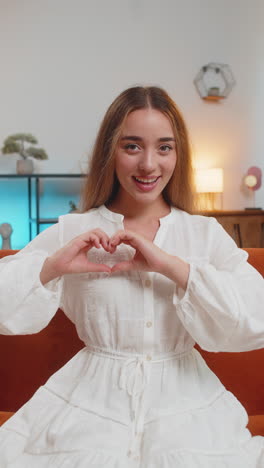 portrait of happy caucasian young woman makes symbol of love showing heart sitting on sofa at home
