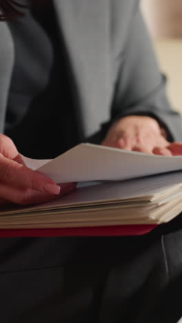 close-up of businesswoman in gray suit flipping through large stack of documents in red folder while sitting in office, showcasing detail-oriented work, organization, and hands-on paper handling process