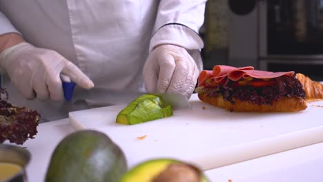 A-chef-cuts-an-avocado-on-a-white-board-with-a-group-of-sandwich-ingredients-around-it