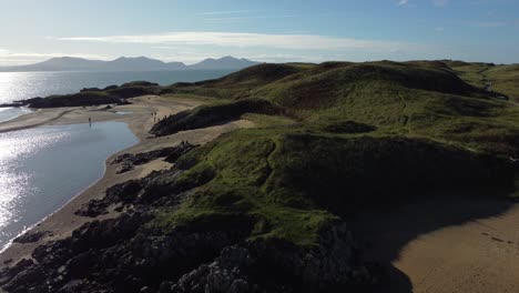 Aerial-rising-view-over-Ynys-Llanddwyn-Welsh-island-with-shimmering-ocean-and-misty-Snowdonia-mountain-range-across-the-sunrise-skyline