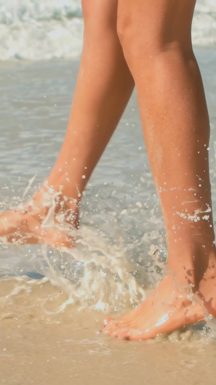 Video verticale: Gambe che camminano lungo la riva in bikini, onde che si infrangono, spruzzi della risacca e impronte.