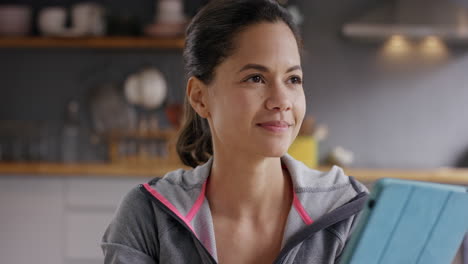 Pretty-mixed-race-woman-using-touch-screen-digital-tablet-at-home-in-kitchen-wearing-sportswear