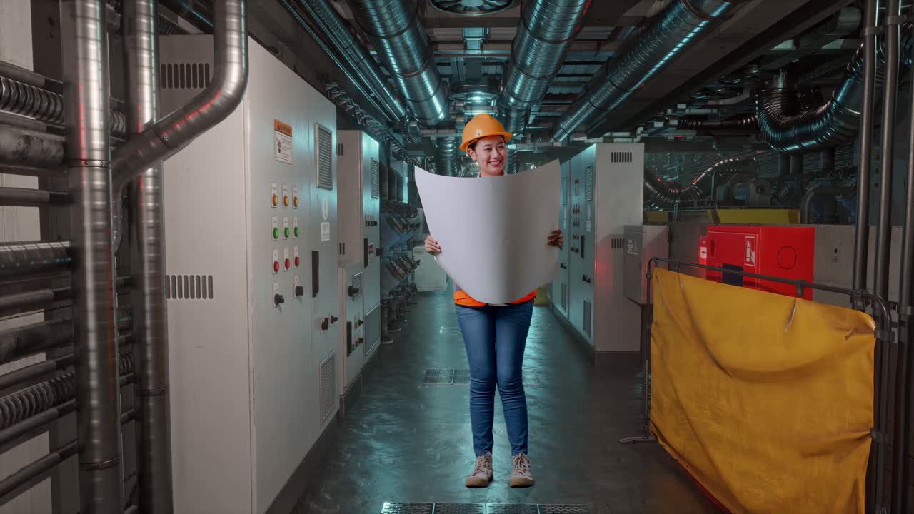 Full Body Of Asian Female Engineer With Safety Helmet Looking At Blueprint In Her Hands And Looking Around While Standing In Engine Control Room, Work Of Electrical Generators