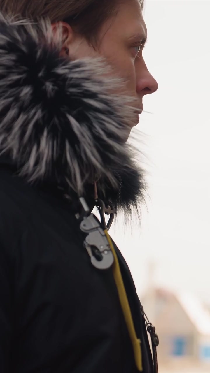 Man outfitted in dark outerwear featuring hooded section. Details include orange stitching near shoulder seam. Environment includes backdrop of hazy atmosphere structural