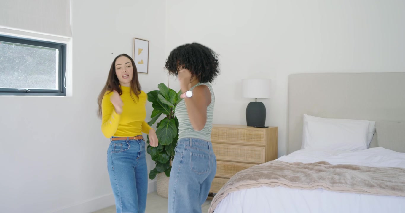 Amigas diversas dançando no quarto depois que a de blusa listrada passa pela cama, enquanto a de blusa amarela se diverte.