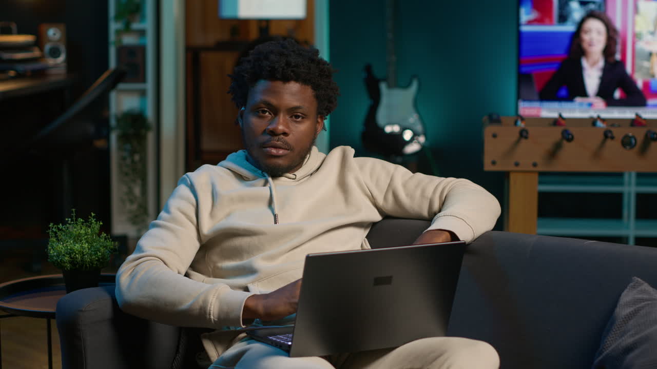 African American freelancer working remotely on a laptop under neon lights, sitting on a couch in a cozy loft apartment. Relaxed man in a stylish home highlights modern productivity. Camera A.