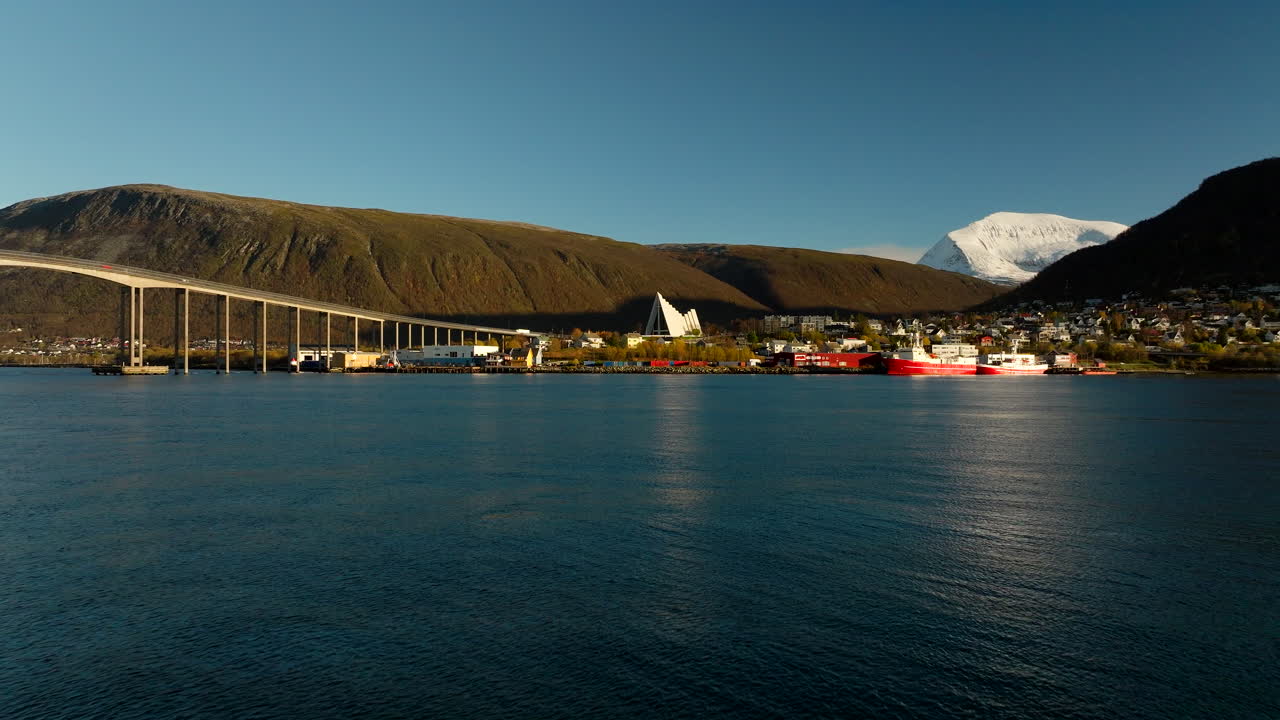 Drone flies toward Arctic Cathedral and Tromso Bridge beneath low sun, Aerial