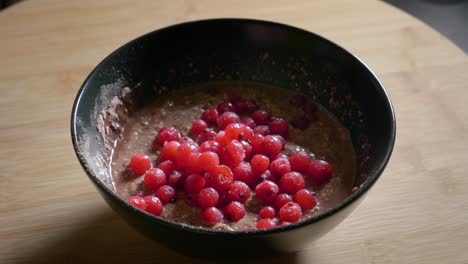 a close up shot tracking in onto a low calorie protein pudding topped with sweet tasty red currants, a delicious healthy dessert served in a black bowl on a wooden kitchen table