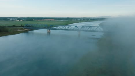 Aerial-establishing-view-of-the-steel-bridge-over-Lielupe-river-on-a-sunny-summer-morning,-fog-rising-over-the-river,-cars-driving,-wide-drone-shot-moving-forward