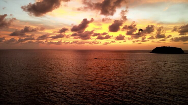 Aerial View Of Parasailer Being Pulled By Boat Against Golden Orange Sunset Skies Over Tropical Waters Off Phuket