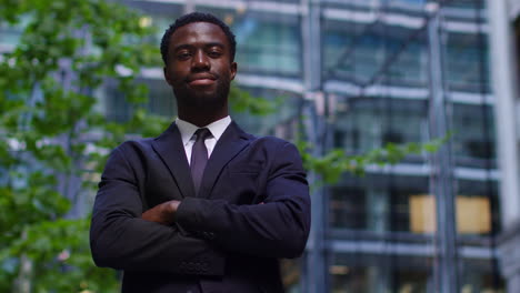 Portrait-Of-Confident-Young-Businessman-Wearing-Suit-Folding-Arms-Standing-Outside-Offices-In-The-Financial-District-Of-The-City-Of-London-UK