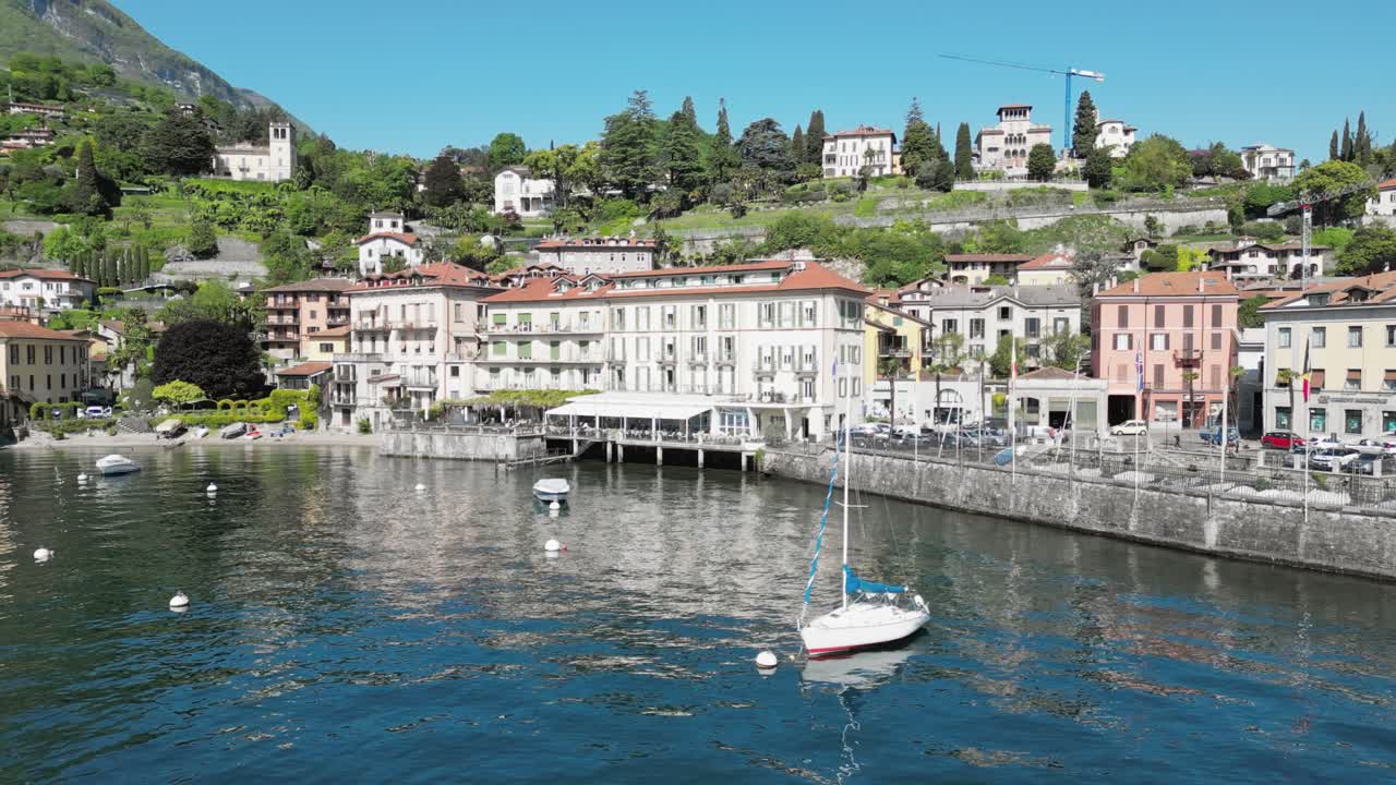 A sailboat and motorboat anchored at a buoy near the lakeside town of Menaggio on Lake Como, surrounded by calm waters and stunning mountain scenery