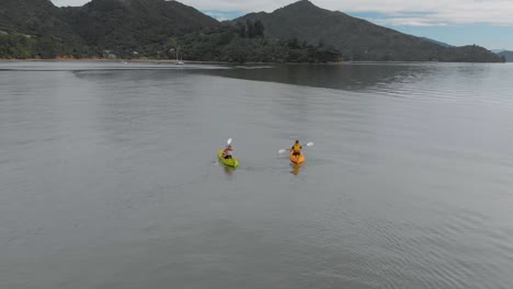 SLOWMO---Two-people-kayaking-with-yachts-in-background-in-Marlborough-Sounds,-New-Zealand---Aerial