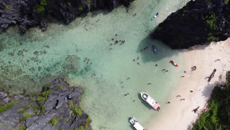 Aerial-spiral-twist-top-down-view-:-Tour-C-Island-Hopping-Boats-docked-on-white-sand-hidden-Beach,-People-swimming-in-tropical-shallow-crystal-clear-lagoon-water-between-jungle-and-Karst-mountains