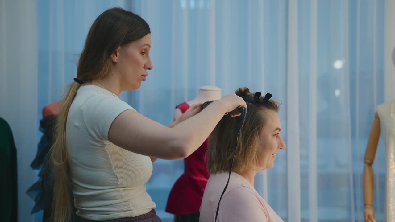 Hairdresser styles client's hair with a curling tool. Client smiles, enjoying the styling session. Studio has mannequins and bright window curtains. Ideal for beauty salons or fashion-related content