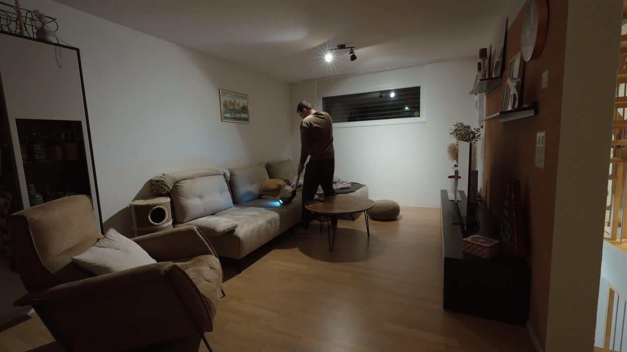 A man vacuums his sofa late at night in a quiet apartment, showing late-hour cleaning, tidying habits, nighttime routine, and focused household maintenance