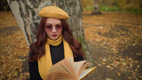 lady with painted lips wearing yellow beret seated outdoors, resting back on tree, flipping through book as she places hand on a particular page, autumn leaves scattered across ground