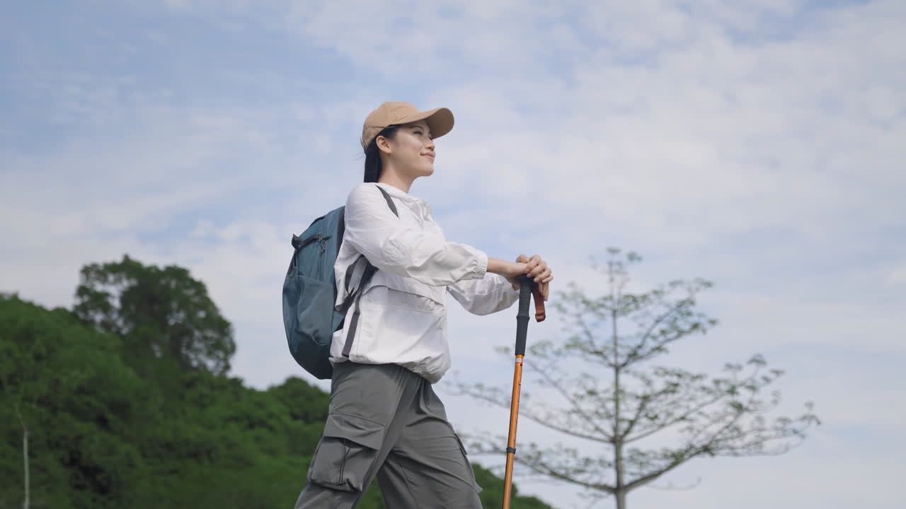Wanita pendaki ransel yang tersenyum dengan ransel dan tongkat trekking, mencapai puncak gunung, beristirahat, menikmati suasana alam dan mengagumi pemandangan setelah berjalan di sepanjang jalur hutan dan mendaki