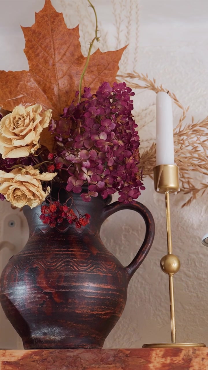 Primo piano di un vaso in ceramica marrone scuro con motivo ondulato. Una candela bianca in un portacandele dorato e un contenitore in ceramica bianca sono presenti. Una piccola zucca rossa con macchie gialle appare parzialmente.