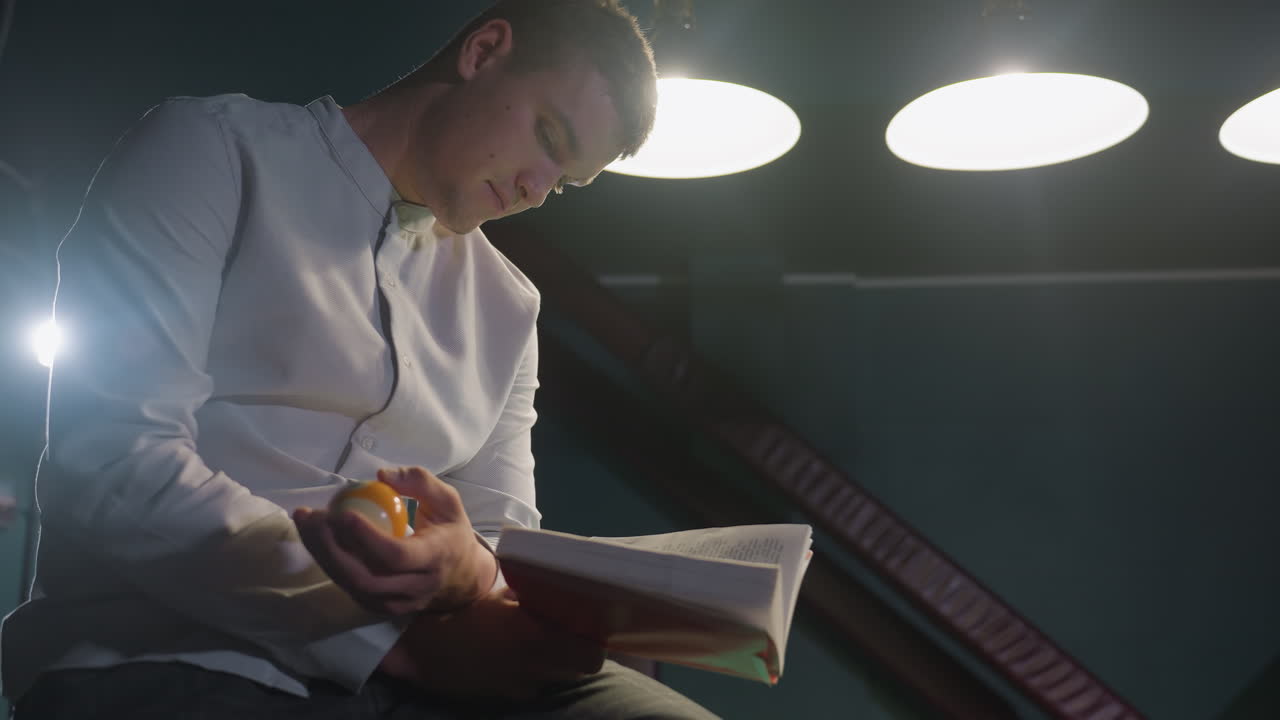 toma de primer plano de un hombre con camisa blanca sentado en una mesa de billar verde, leyendo un libro mientras tira una pelota de billar en la mano. expresión enfocada, iluminación de mal humor y fondo dramático