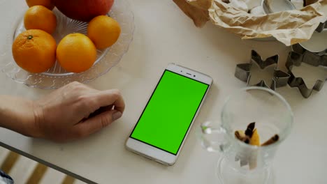 primer plano de la mano de la mujer navegando por el teléfono inteligente con pantalla verde en la mesa de la cocina en casa