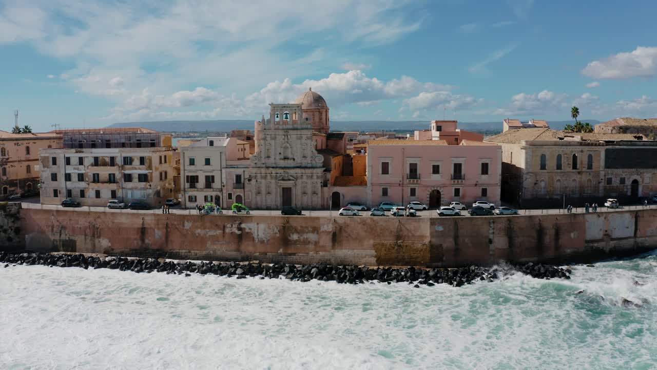 Aerial drone shot of historic buildings on the island of Ortigia with waves crashing on the rocks. Medieval town in Sicily with natural fortress wall along the coast in Sicily. Old town of Syracuse.