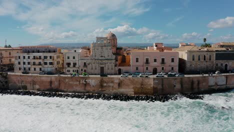 Aerial drone shot of historic buildings on the island of Ortigia with waves crashing on the rocks. Medieval town in Sicily with natural fortress wall along the coast in Sicily. Old town of Syracuse.