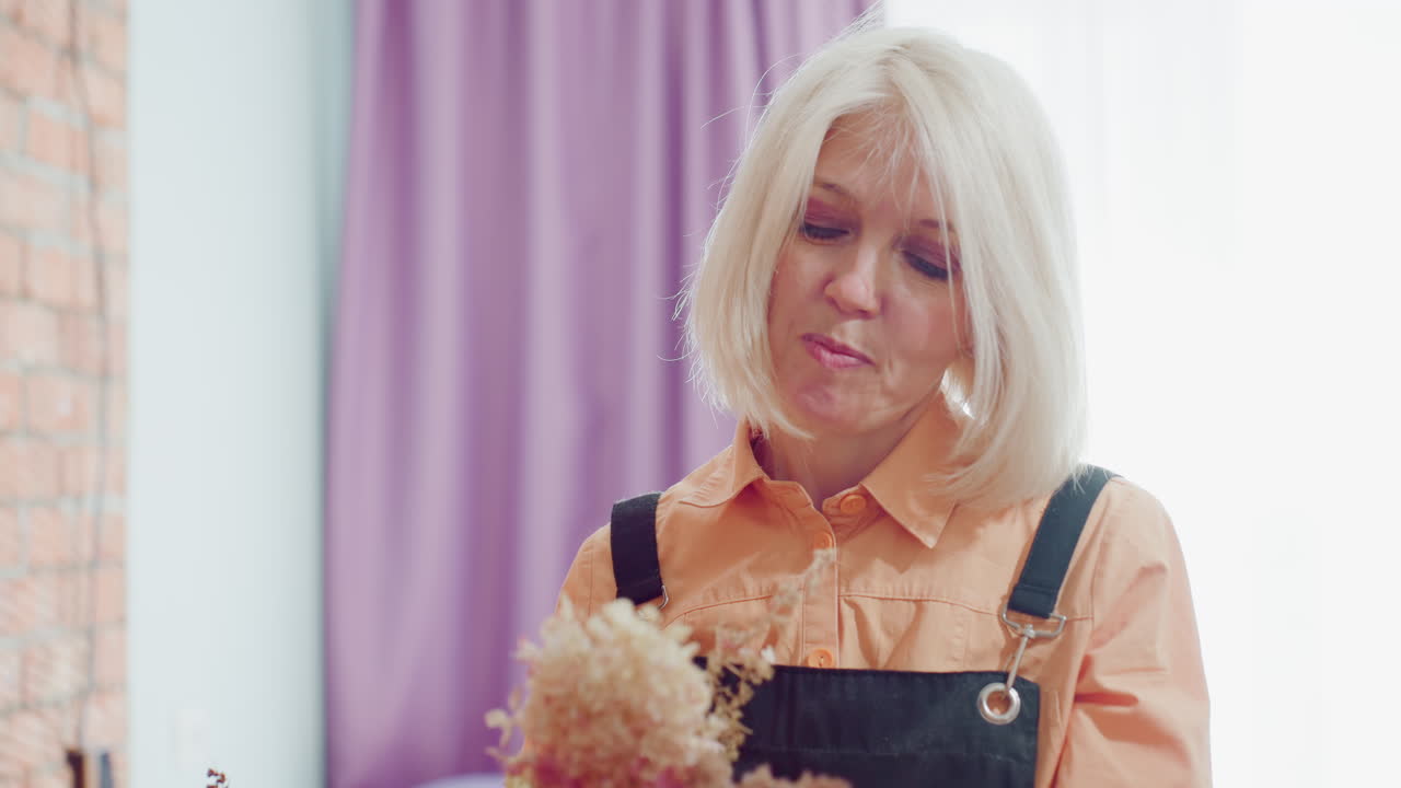 Florist in black apron and orange shirt arranges delicate dried hydrangea flowers in glass vase with gentle hands, working with focus and care in bright workshop decorated with purple curtains