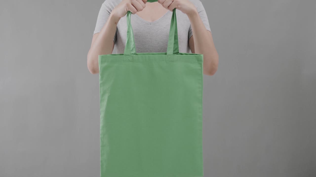 mujer caucásica con una camiseta blanca sosteniendo una bolsa verde sobre un fondo gris, espacio de copia, cámara lenta.