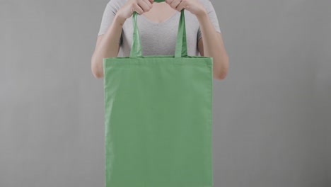 mujer caucásica con una camiseta blanca sosteniendo una bolsa verde sobre un fondo gris, espacio de copia, cámara lenta.