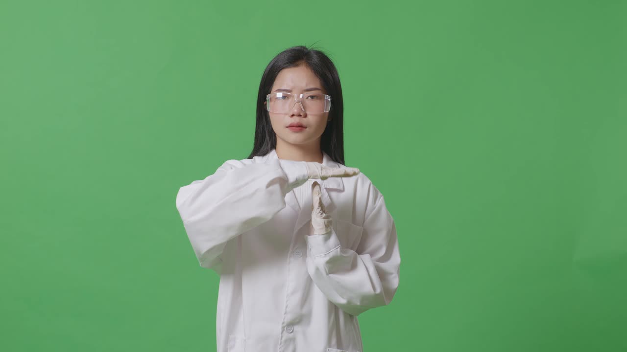 화가 난 아시아 여성 과학자는 머리를 흔들고 실험실의 녹색 스크린 배경에 서서 손으로 타임 아웃 제스처를 하고 있습니다.