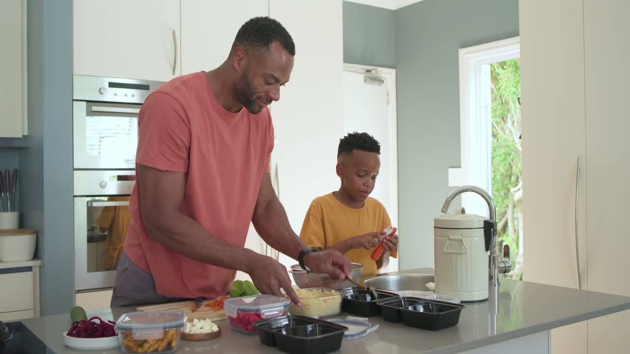 Padre e figlio afroamericani con magliette color salmone e senape, dopo la preparazione, riempiono vassoi neri per pasti all'isola della cucina