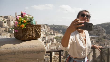 beautiful elegant young woman in matera italy taking selfies pictures with smartphone sitting on a high wall viewpoint with landscape. fashion dress with hat and skirt.