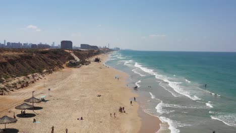 Immer-Mehr-Touristen-Liegen-Gerne-Im-Schatten-Eines-Strohgedeckten-Sonnenschirms,-Während-Andere-Touristen-An-Einem-Heißen-Sommertag-Am-Strand-Von-Herzeliya-In-Israel-Das-Klare-Blaue-Mittelmeer-Genießen