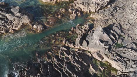 Low-overhead-backwards-flight-over-granite-rocky-shoreline-with-crystal-clear-water-on-a-sunny-day