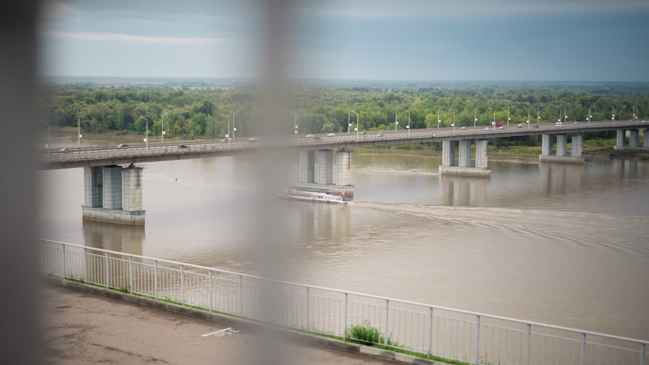 흐릿한 난간이 비 오는 강 다리 프레임을 형성하고, 작은 배가 콘크리트 기둥 아래 흙탕물을 지나가요. 흐린 하늘, 멀리 보이는 수목선, 얕은 심도가 사색적인 영화 같은 분위기를 연출하는 도시 풍경입니다.