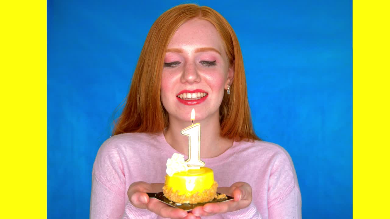 한 젊은 여성이 파란색과 노란색을 배경으로 밝은 스튜디오에서 미소를 지으며 1번 모양의 양초가 달린 작은 케이크를 들고 있습니다.