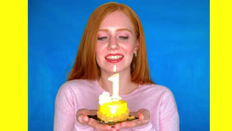 a young woman stands smiling in a bright studio against a blue and yellow backdrop, and holds a small cake with a number one shaped candle