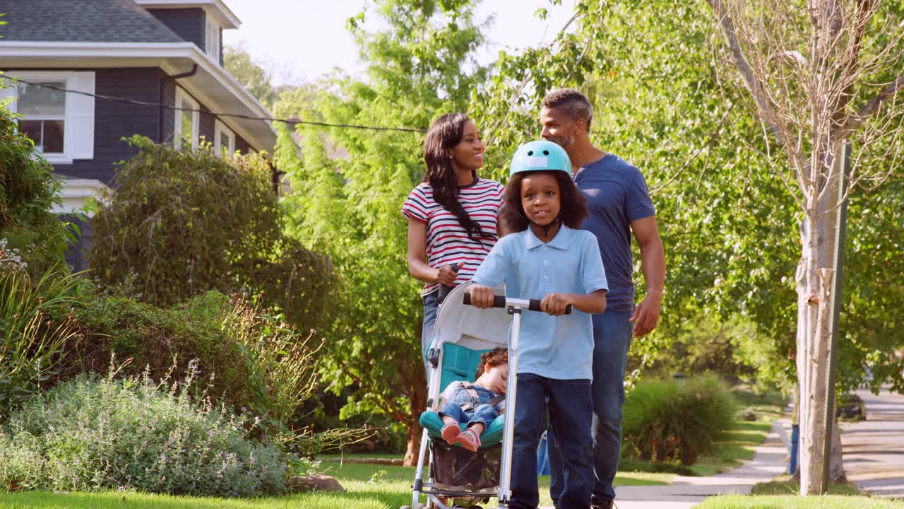 una pareja empuja a su hija en un cochecito mientras su hijo monta un scooter.