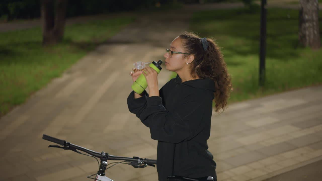 Athletic Woman Drinks After Cycling Session, Active Woman Takes Large Gulp From Green Bottle After Ride, Focused Athlete Pauses To Hydrate From Green Container Following Evening Cycling Workout