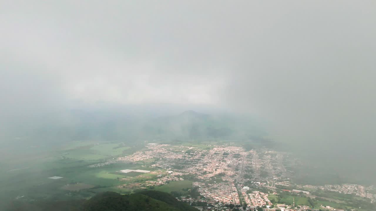 Small Mexican town surrounded by farmland slowly emerges through dense clouds near Tuxpan, Mexico on a rainy day. Slow motion drone aerial