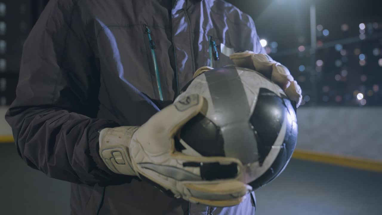 primer plano de manos con guantes frotando una pelota de fútbol desgastada durante una intensa sesión de entrenamiento nocturno en un campo urbano, luces brillantes en el fondo