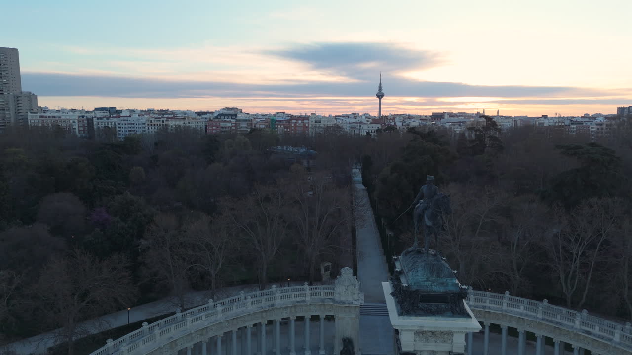 Drone flies over Retiro Park in Madrid, showcasing the Alfonso XII monument, the lake, and a stunning city skyline at sunset. The golden hour light enhances the breathtaking urban and natural scenery