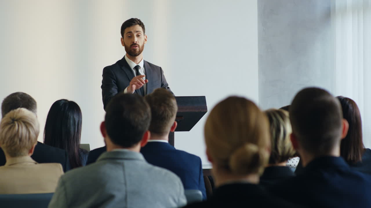 Caucasian businessman speaker on a podium wearing formal clothes and talking in a conference room in front to many people