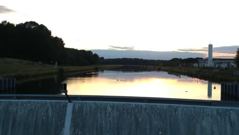 drone pans over the edge of a damn while birds fly over the reflection of the sunset