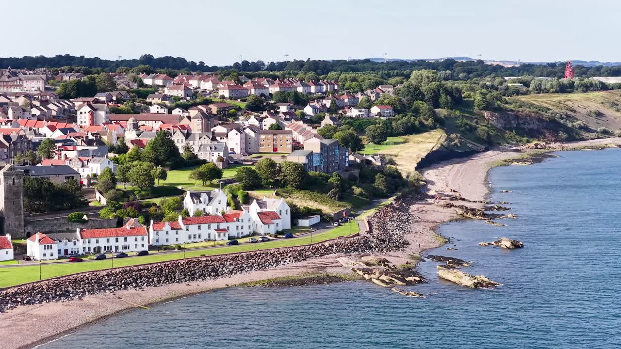 Drone glides above seaside village, revealing houses, rocky shoreline, and lush landscape in daylight