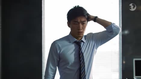 a young man in a blue dress shirt stands in his office, looking perplexed and stressed. his hand is on his head as he faces an apparent dilemma.
