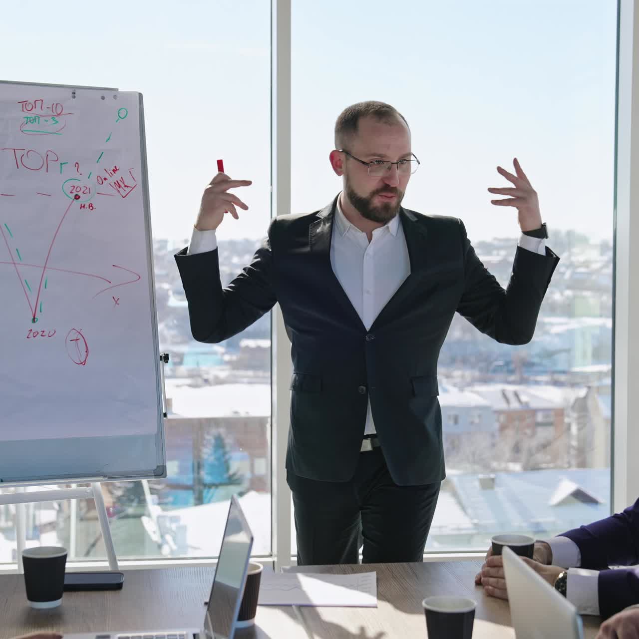 Charismatic speaker explaining something to the colleagues in front of a chart. Male speaker giving a speech smiling and gesturing actively. Cityscape at the background